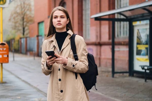 Atrakcyjne stylowa dziewczyna z plecakiem starannie za pomocą telefonu komórkowego czeka na transport publiczny na przystanku autobusowym na zewnątrz — Zdjęcie stockowe