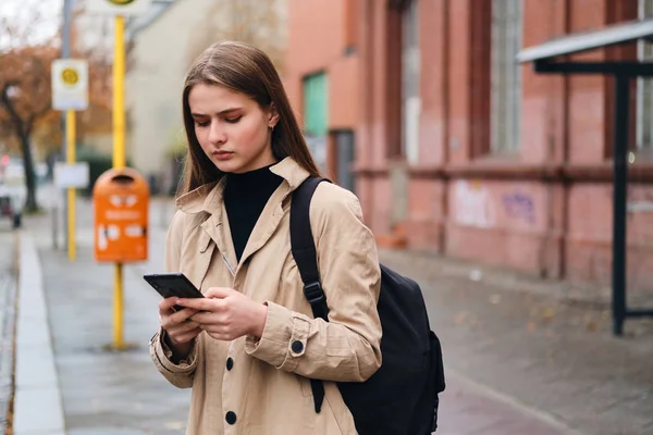 Seriøs, stilig jente med ryggsekk som venter på bussholdeplass utendørs med mobiltelefon – stockfoto