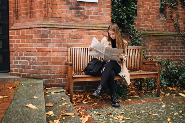 Привлекательная случайная девушка с рюкзаком вдумчиво читает газету на скамейке под открытым небом — стоковое фото
