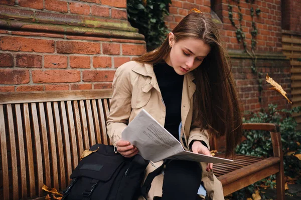 Atraktivní ležérní dívka zamyšleně číst noviny sedí s batohem na lavičce venku — Stock fotografie