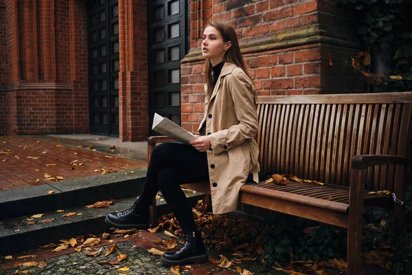 Piękna stylowa dziewczyna z pewnością patrząc daleko czytając gazetę na ławce outdoor — Zdjęcie stockowe