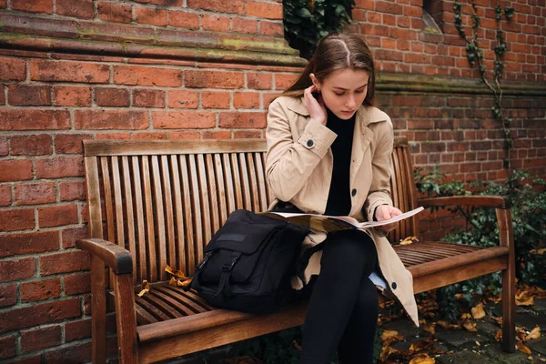 Krásná stylová dívka soustředěně čtení novin odpočívá na lavičce venku — Stock fotografie