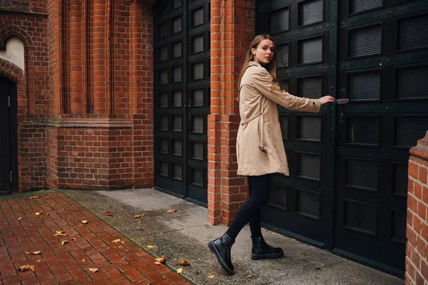 Hermosa chica casual en gabardina mirando atentamente en la cámara abriendo la puerta vieja en la calle — Foto de Stock