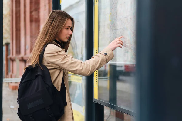 Gadis santai yang cantik dengan ransel yang tampak seperti peta kota di halte bus di luar ruangan — Stok Foto