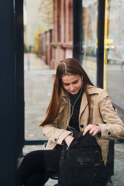 Attraktiv, tilfeldig jente i øretelefoner med ryggsekk ventende på offentlig transport på bussholdeplassen utendørs – stockfoto