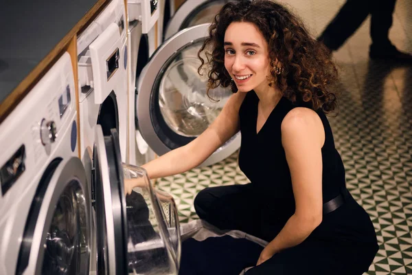 Bella ragazza bruna felicemente alla ricerca in macchina fotografica togliendo i vestiti dalla lavatrice in lavanderia self-service — Foto Stock