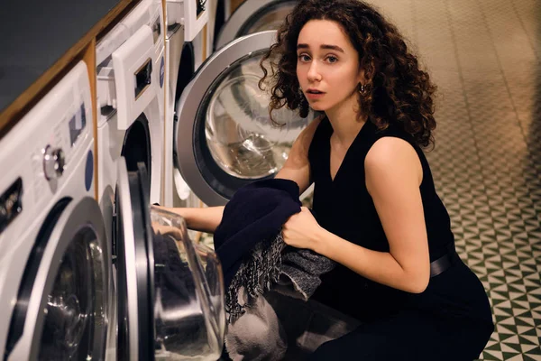 Retrato de chica morena mirando pensativamente en la cámara sacando la ropa de la lavadora en la lavandería de autoservicio — Foto de Stock