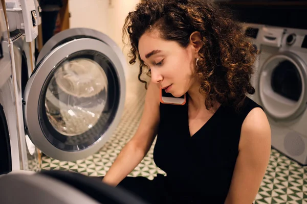 Bella ragazza bruna che parla su smartphone lavando i vestiti nella moderna lavanderia self-service — Foto Stock