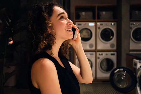 Vista laterale di bella ragazza felicemente parlando su smartphone in attesa di lavaggio in lavanderia self-service di notte — Foto Stock