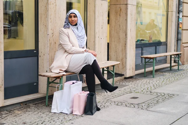 Young beautiful Arabic woman in hijab resting on bench with shopping bags on city street