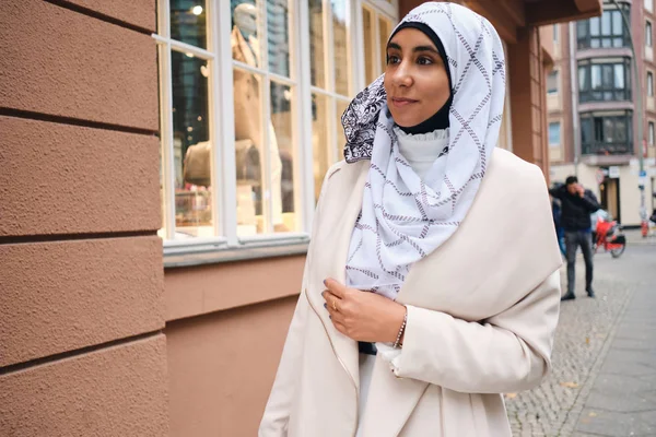 Young attractive Arabic woman in hijab dreamily walking through city street