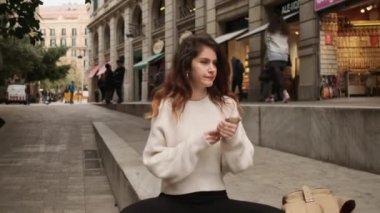 Akıllı telefonlu çekici İspanyol bir kızın şehir caddesinde oturan birini çekimini izliyoruz.