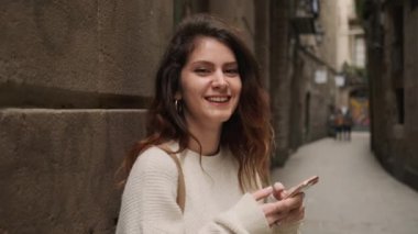 Çekici İspanyol tebessümlü kız kameraya mutlu bir şekilde bakıyor dar şehir caddesinde akıllı telefon kullanıyor.