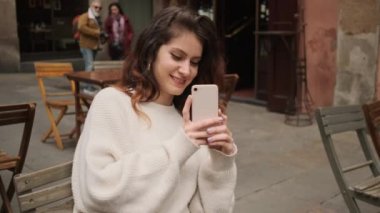 Güzel, neşeli İspanyol kız dışarıda dinlenen bir kafede akıllı telefondan fotoğraf çekiyor.