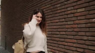 Sırt çantalı ve akıllı telefonlu çekici bir kız şehrin sokaklarında tuğla duvar boyunca mutlu mesut yürüyor.
