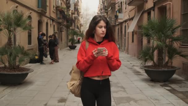 穿着风衣背着背包带着智能手机穿过旧城区街道的惊慌失措女孩的追踪镜头 — 图库视频影像