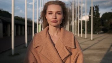 Paltolu güzel sarışın kız sokakta kameraya bakıyor. Gün batımında dışarıda poz verin.