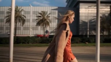 Paltolu ve ipek elbiseli seksi sarışının kavramsal karesi gün batımında güvenli bir şekilde caddede yürüyor.