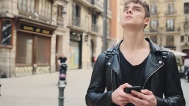 Deri ceketli, akıllı telefonlu, yakışıklı bir gencin caddeye dikkatlice baktığı bir fotoğrafı.