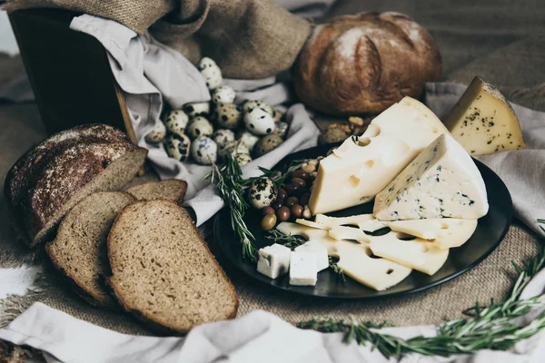 Cheeses lying on black dish and bread situated nearby. Blue cheese, cheese with holes decorated with herbs. Big piece of round bread and tasty slices ready for making sandwiches for a snack. — Stock Photo, Image
