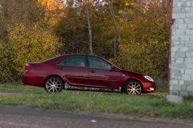 Kiraz kırmızı 4 kapılı aile d-sınıfı sedan Toyota Camry beşinci nesil xv30 2001-2006 led farlar ve 18 inç jantlar büyük jantlar ile yaz bir arka plan üzerinde yeşil bir alan ve orman üzerinde ayakta