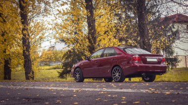 Kiraz kırmızısı 4 kapılı sonbahar sahnesi d sınıfı sedan Toyota Camry beşinci nesil XV30 2001-2006 Yazın yeşil bir tarla ve arka planda duran büyük farlar ve 18 inç jantlarla