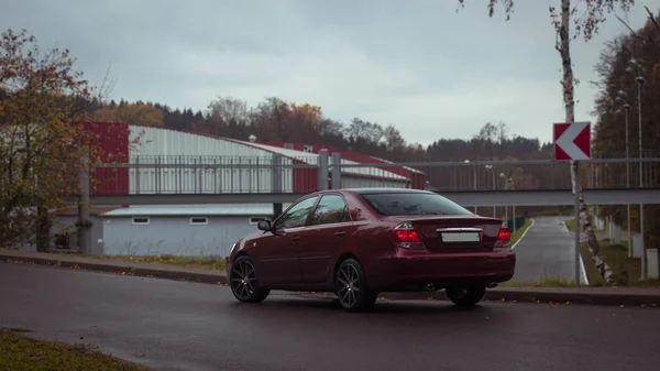 Kiraz kırmızısı 4 kapılı sonbahar sahnesi d sınıfı sedan Toyota Camry beşinci nesil XV30 2001-2006 Yazın yeşil bir tarla ve arka planda duran büyük farlar ve 18 inç jantlarla