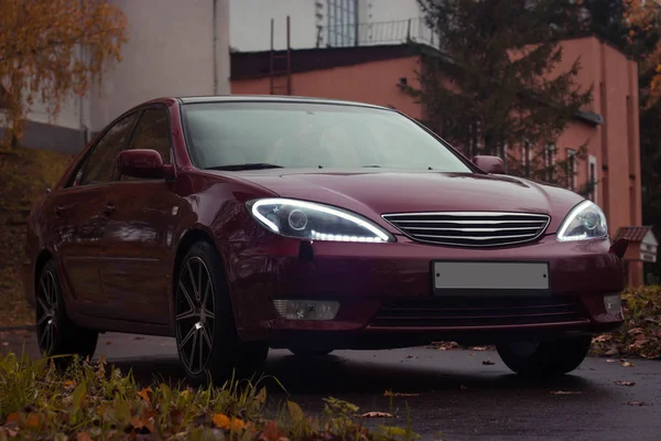 Kiraz kırmızısı 4 kapılı sonbahar sahnesi d sınıfı sedan Toyota Camry beşinci nesil XV30 2001-2006 Yazın yeşil bir tarla ve arka planda duran büyük farlar ve 18 inç jantlarla