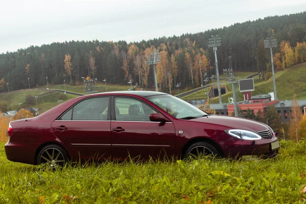 Kiraz kırmızısı 4 kapılı sonbahar sahnesi d sınıfı sedan Toyota Camry beşinci nesil XV30 2001-2006 Yazın yeşil bir tarla ve arka planda duran büyük farlar ve 18 inç jantlarla