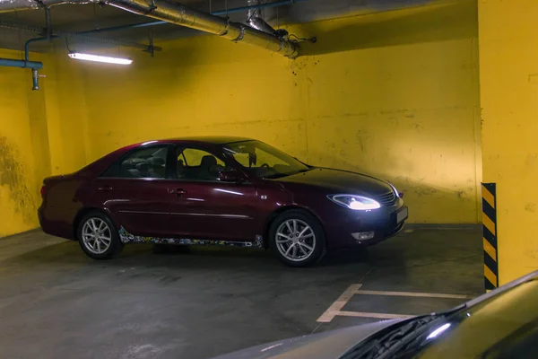 Vişne kırmızısı 4 kapılı yeraltı otoparkı D sınıfı sedan Toyota Camry beşinci nesil XV30 2001-2006 gri bir tarlada duruyor ve arka planda sarı duvarlar kurşun farları ve 16 inç jantları var. 