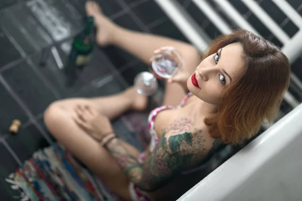 Beautiful girl with colorful tattoos sits near white bath on the — Stock Photo, Image