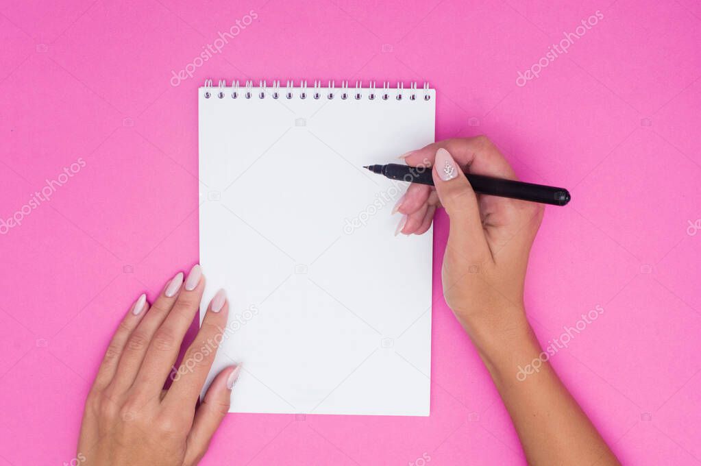 Notebook with pen holds female hands on a pink background.