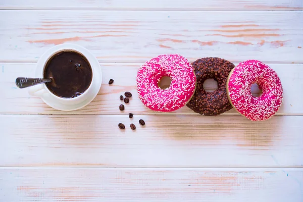 Вкусные и вкусные пончики с розовой глазурью и порошок с чашкой ароматного кофе на белом деревянном фоне . — стоковое фото