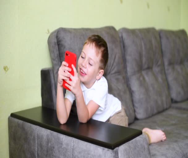 El niño se sienta en un sofá gris y habla por teléfono. Un joven bloguero . — Vídeo de stock