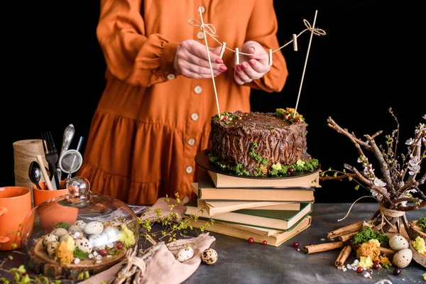 Beautiful composition with cake, books, cups, eggs, flowers, twigs. The girl attaches a thread to the cake for attaching photos.