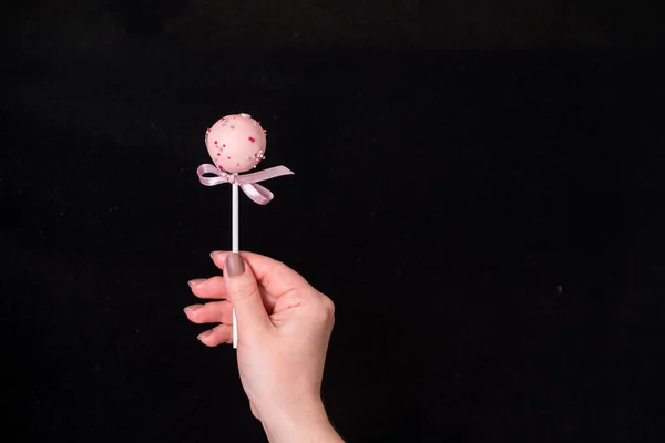Cake pops in women\'s hands on a black background. Dessert in pink chocolate cream with powder and bow. Tasty food