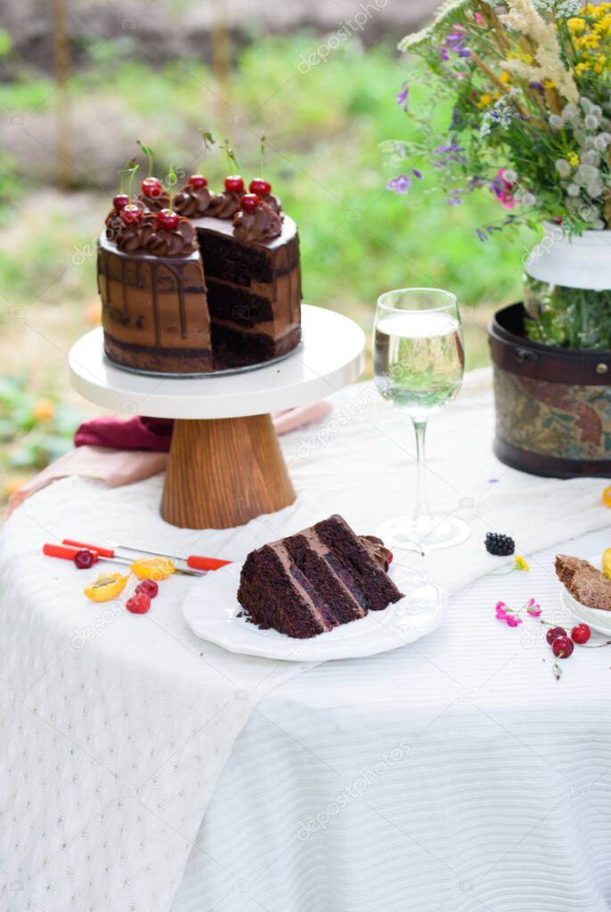 Delicious chocolate cake decorated with cherries with a glass of white wine, berries and wild flowers. Picnic in the country