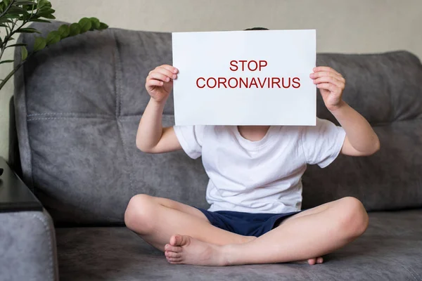Chłopiec siedzi na kanapie z białą kartką papieru przed twarzą z napisem stop coronavirus, i zakrywa twarz. — Zdjęcie stockowe