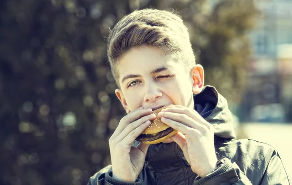 Chico joven comiendo una hamburguesa con queso en la naturaleza —  Fotos de Stock