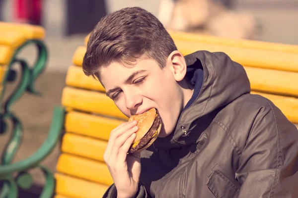 Chico joven comiendo una hamburguesa con queso en la naturaleza —  Fotos de Stock