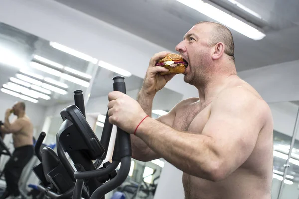 Man eats a hamburger with meat and cheese in the gym — Stock Photo, Image