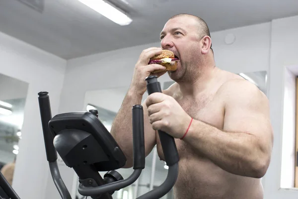 man eats a hamburger with meat and cheese in the gym