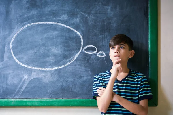 Student chłopiec, opierając się na tablicy i myślenia w szkole — Zdjęcie stockowe