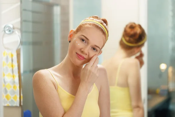 Mujer joven aplica crema anti edad en la cara sonriendo — Foto de Stock