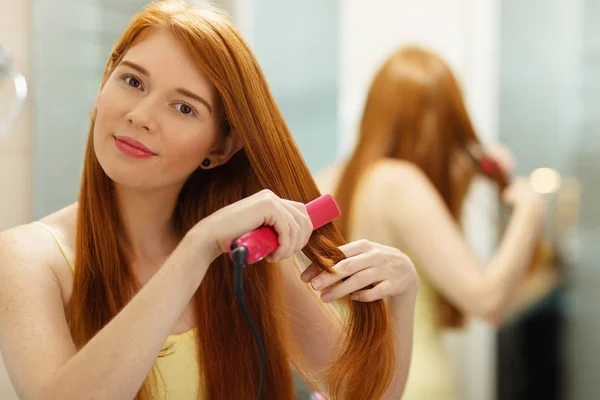 Bella donna stiratura sana capelli rossi con ferro raddrizzatore — Foto Stock