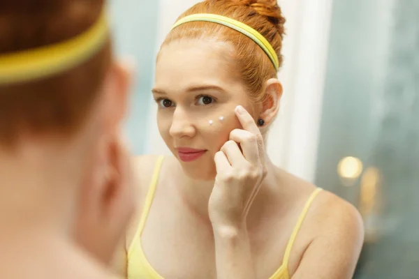 Mujer joven aplica crema anti-envejecimiento mirando espejo — Foto de Stock