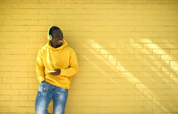 Joven africano escuchando música apoyada en la pared amarilla —  Fotos de Stock