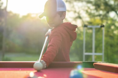 Küçük bir çocuk açık havada bilardo oynuyor.