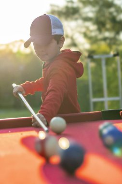 Küçük bir çocuk açık havada bilardo oynuyor.