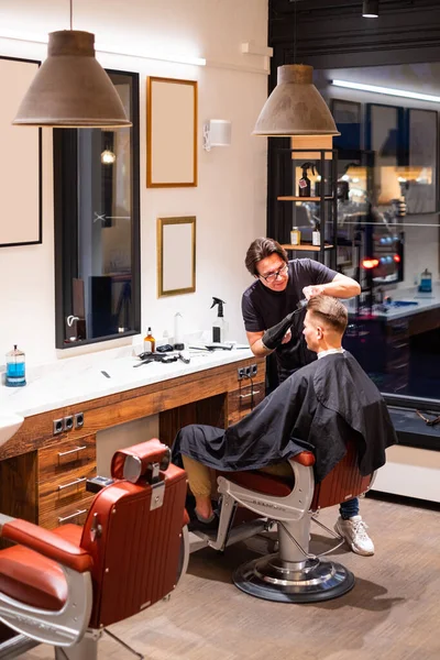 男理发师用吹风机擦干一个年轻人的头发 并用刷子在理发店里给他梳头 — 图库照片